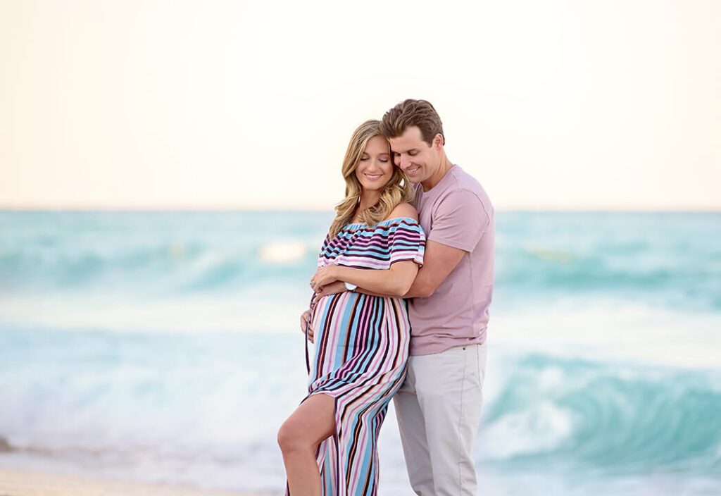 Erika Rakow and Scott embracing on a sandy beach, showcasing their beach maternity portraits in Palm Beach County.