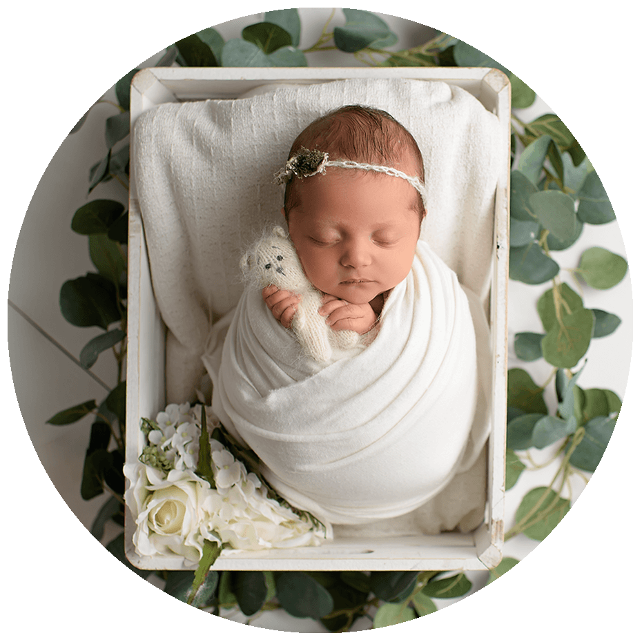 Newborn and Infant Photography in Jupiter Florida; Cute baby girl with headband accessory wrapped in a newborn wrap holding a stuffed bear and surrounded by green leaves.